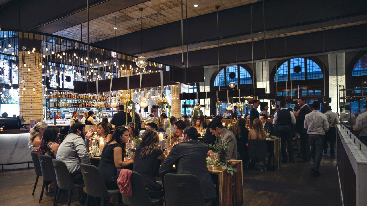 Upscale restaurant filled with guests at long wooden tables under warm string lights and hanging bulbs.