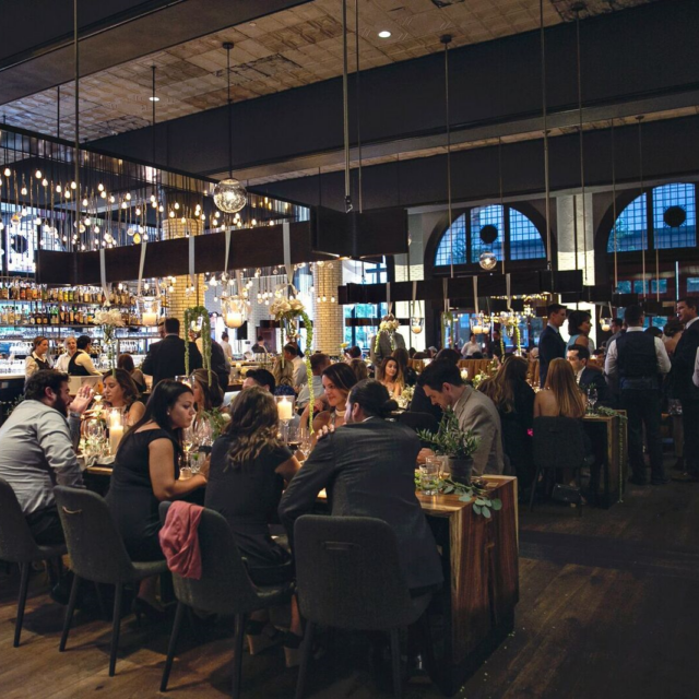 Upscale restaurant filled with guests at long wooden tables under warm string lights and hanging bulbs.