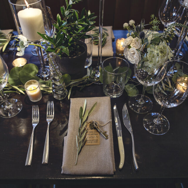Elegant dining table set with candles, glassware, and greenery, featuring a beige napkin and sprig at each place setting.