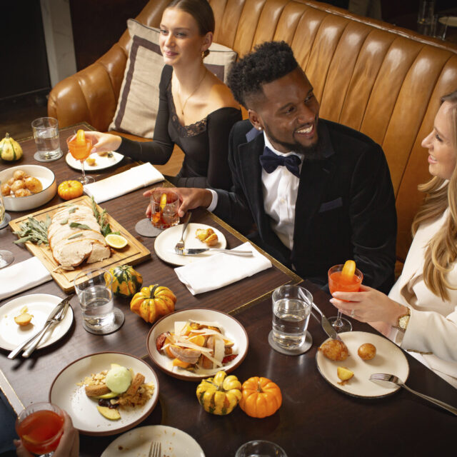 People seated around a long table with autumn decor and small pumpkins, enjoying a meal and drinks in a restaurant setting.
