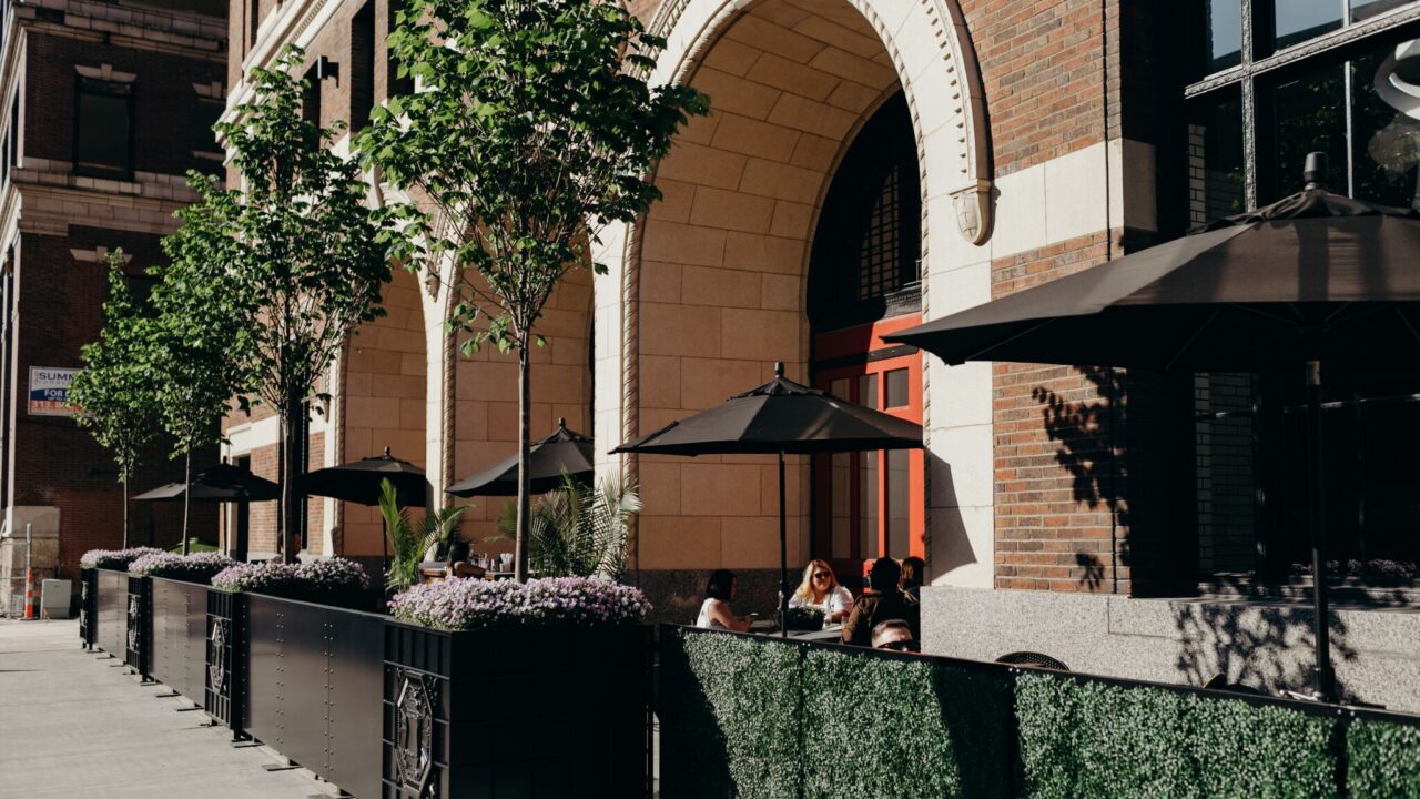 Outdoor cafe seating beside a brick building with a large arched entrance, black umbrellas, planters, and green hedges.